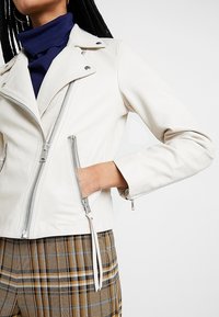 Bílá kožená biker bunda se stříbrnými zipy, přiléhavý střih a výrazný klopový límec. Kombinováno s károvanými kalhotami v hnědo-modrém vzoru.