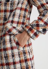 Main dans la poche d'un manteau en laine à carreaux blancs, rouges, noirs et moutarde avec des boutons noirs et un tissu texturé.