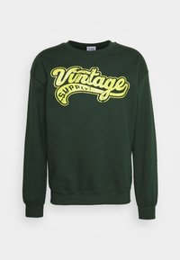 Zelená mikina se širokým výstřihem, dlouhými rukávy a žlutým vintage-style grafickým nápisem "Vintage Supply" vpředu.