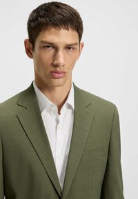 Grüner strukturierter Blazer mit taillierter Jackenform, einer Brusttasche, getragen über einem weißen gemusterten Hemd mit Knopfdetails.
