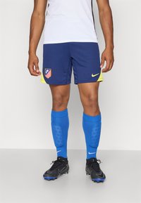 Nike sports shorts i marineblå med gule detaljer, der viser et holdlogo. Blå fodboldsokker, sorte støvler med grå og blå detaljer.