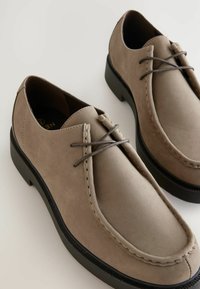 Pantofi din suede bej cu vârf rotunjit, detalii cusute și șireturi gri. Talpa din cauciuc negru oferă un contrast față de materialul superior.