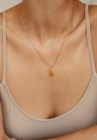 Collana d'oro con un pendente rettangolare, caratterizzato da una sfumatura giallo-arancio, indossata sopra un top in beige, che mette in risalto il décolleté.