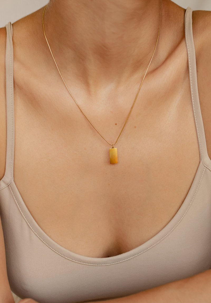 Collana d'oro con un pendente rettangolare, caratterizzato da una sfumatura giallo-arancio, indossata sopra un top in beige, che mette in risalto il décolleté.