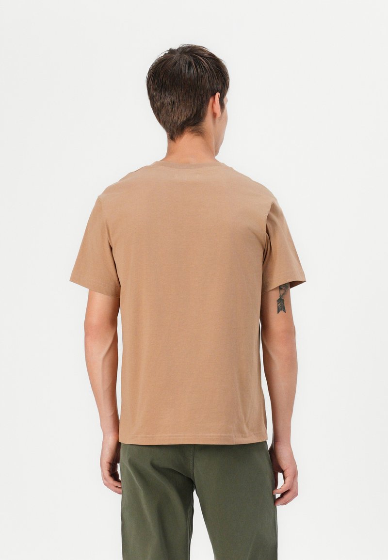 T-shirt in cotone marrone con maniche corte e colletto rotondo, caratterizzato da una vestibilità rilassata e dettagli di cucitura minimali, indossato con pantaloni verdi.