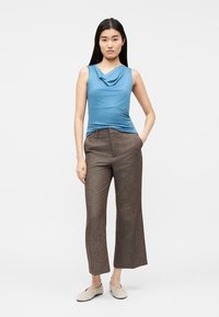 Γυναίκα στέκεται με το ένα χέρι στην τσέπη, φοράει μπλε αμάνικο τοπ με καπελούδα λαιμόκοψη, καφέ παντελόνι φαρδύ και κομμένο στο μήκος, και μπεζ επίπεδα παπούτσια.