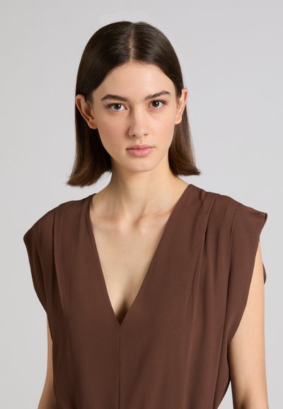 Junge Frau mit glattem braunem Haar, die ein ärmelloses braunes Top mit tiefem V-Ausschnitt trägt, schaut nach vorne vor einem schlichten grauen Hintergrund.