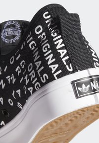 Svarta sneakers med ett texturerat tygöverdel som har vit tryckt text. Vit gummisula med ett mönstrat botten och varumärkesdetaljer.