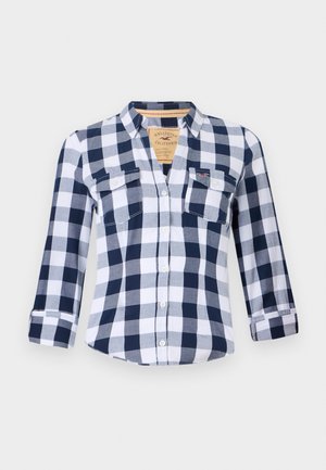 Chemise à manches longues à carreaux bleu marine et blanc, avec deux poches avant, design boutonné et ourlet arrondi. Le tissu semble doux et lisse.