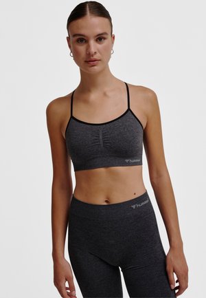 Kvinna som bär en mörkgrå sömlös sport-bh och leggings, stående mot en enkel ljus bakgrund, vänd framåt med avslappnade armar.