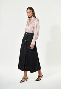 Blusa di raso rosa chiaro con colletto arricciato, abbinata a una gonna in denim scuro a vita alta con bottoni dorati e tasche a toppa. Sandali neri con tacco.