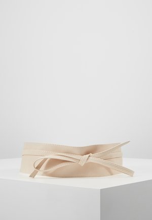 Beige bredt bælte med en lang tynd rem bundet i en sløjfe, udstillet på en hvid piedestal mod en ensfarvet baggrund.