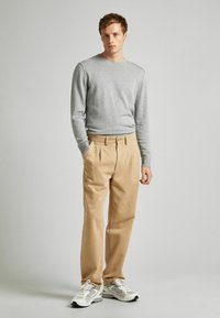Helles graues Sweatshirt mit gerippten Bündchen, kombiniert mit beigen, plissierten Hosen und weißen Sneakers mit grauen Akzenten. Neutraler Hintergrund.