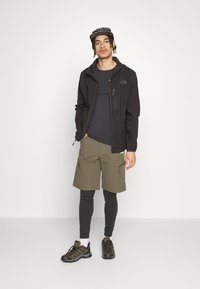 Czarna kurtka zapinana na zamek na ciemnoszarym T-shircie, połączona z oliwkowymi szortami i czarnymi legginsami, uzupełniona brązowym obuwiem wędrownym.
