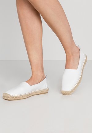 Parbare ben iført hvide espadrille slip-on sko med vævede jutebund på en ensfarvet baggrund.