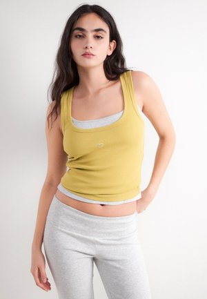 Jeune femme aux longs cheveux foncés portant un débardeur jaune par-dessus un sous-vêtement gris et un pantalon gris clair, debout devant un fond blanc.