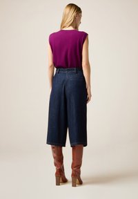 Top morado sin mangas combinado con culottes de denim oscuro de tiro alto; botas de cuero marrón hasta la rodilla con tacón ancho completan el atuendo.