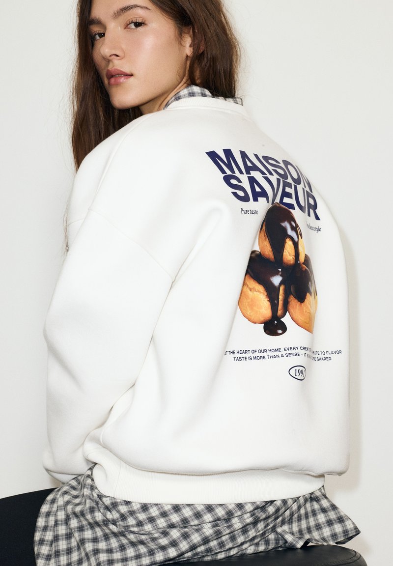 Felpa bianca con una grafica di un dolce con cioccolato che cola, decorata con testo. Indossata sopra una camicia a quadri.