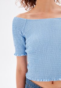 Světle modrý top bez ramínek se smocked texturou, krátkými nadýchanými rukávy a volánkovým lemem. Párováno s džíny.