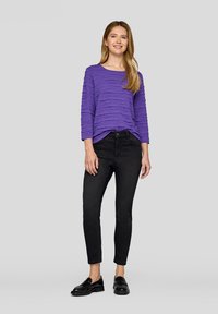 Lila strukturierte Bluse mit horizontalen Rüschen, kombiniert mit schwarzen schmal geschnittenen Jeans und schwarzen Slippers. Das Modell steht vor einem schlichten Hintergrund.