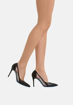 Gambe che indossano calze velate color carne e décolleté nere con punta affilata e pannelli laterali in rete trasparente, in piedi su sfondo bianco.