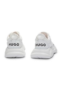 Valkeat urheilukengät, joissa on teksturoidulla verkkomateriaalilla varustettu päällinen, paksu kumipohja ja mustat "HUGO"-brändäykset kantapäässä sekä sivupaneeleissa.