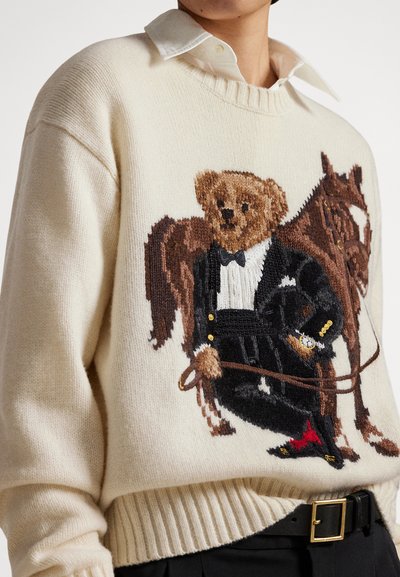 Osoba w kremowym swetrze z dzierganym wzorem misia w fraku trzymającego wodze konia stojąca obok brązowego konia.