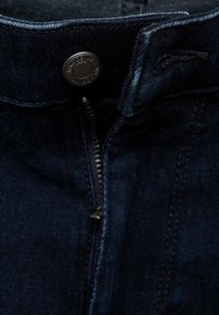 Tela de mezclilla azul oscuro con cremallera visible y botón de metal negro etiquetado "MNG DENIM" en la cinturilla.