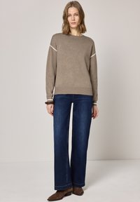 Femme debout portant un pull taupe, un jean large bleu foncé, des bottines marron et un bracelet foncé au poignet gauche.