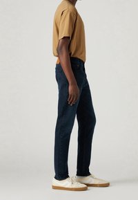 Osoba v hnědém tričku, tmavě modrých slim-fit džínách a béžových teniskách stojící bokem k jednobarevnému pozadí.
