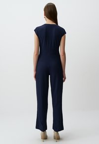 Combinaison bleu marine avec des manches courtes, en tissu texturé, taille cintrée et pantalons à jambe large. Portée avec des escarpins beiges.