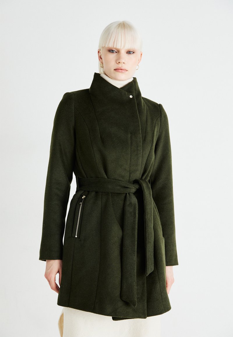 Cappotto verde in lana con colletto alto, cintura a cravatta e tasche laterali con zip. Presenta spalle strutturate e una texture liscia.