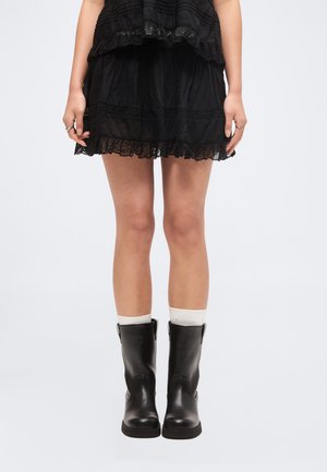 Žena nosi čiernu sukňu s čipkovým lemom, biele ponožky a čierne kožené topánky do polovice lýtok, stojí pred jednoduchým svetlým pozadím.