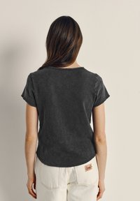 Ženska z dolgimi rjavimi lasmi, oblečena v izprano črno kratko majico in bele hlače z majhno značko na zadnjem žepu, obrnjena stran.