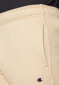 Beige sweatpants with a soft texture, featuring a side pocket and a small embroidered logo in black, white, and red near the hem.