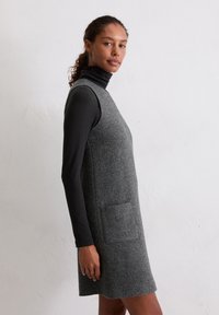 Grijze wollen jurk met een mouwloos ontwerp, met een gestructureerde afwerking en een voorzak. Gedeeltelijk gedragen over een zwart langemouwen top.