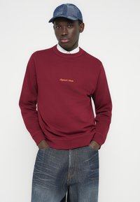 Tamsiai raudonas džemperis su apvalia iškirpte ir ilgomis rankovėmis, dekoruotas auksiniu siuvinėtu tekstu. Derinamas su mėlynais džinsais, kuriuose yra kontrastingos siūlės.