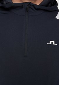 Marintröja med huva och kvartsdragkedja, tillverkad av ett slätt tyg. Har en vit logotyp på bröstet och texturerade detaljer vid halslinningen.