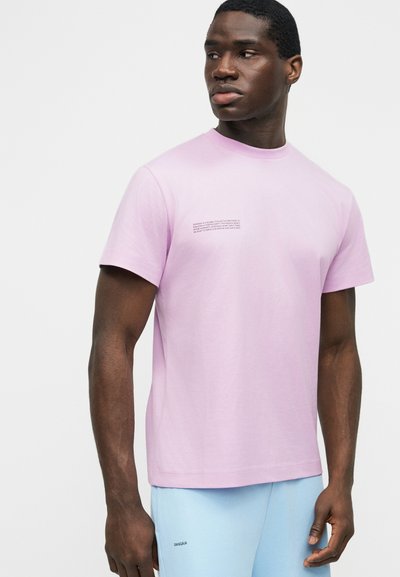 Άνδρας που φοράει ανοιχτό ροζ πουκάμισο με κοντά μανίκια και μικρό μαύρο κείμενο στο στήθος, και ανοιχτό μπλε παντελόνι με το λογότυπο «PANGAIA» στο μηρό.