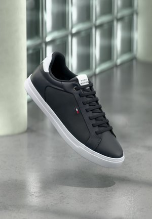 Black low-top sneaker with white sole and heel tab, black laces, and small Tommy Hilfiger logo on side, positioned in a modern indoor setting.