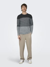 Suéter de manga larga a rayas grises con un degradado de gris oscuro en los hombros a gris claro en la cintura, combinado con pantalones beige de corte holgado y zapatillas blancas.