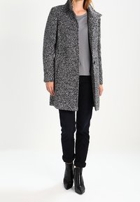 Grå og sort tweedfrakke med krave, med lange ærmer og to forlommer, kombineret med en grå skjorte og sorte jeans.