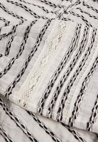 Bílá tkanina s černými pruhovanými vzory a texturovaným krajkovým lemem, s tkanými liniemi a složitými detaily po celém povrchu.
