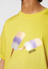 Camiseta de algodón de color amarillo brillante con un gráfico de dos paletas de colores y un degradado en púrpura claro, además del logo de MCM en negro.