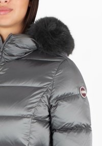 Giacca a vento grigia con cappuccio, dotata di un pom-pom in pelliccia sintetica. Il materiale è lucido con sezioni trapuntate e una patch con logo sulla manica.