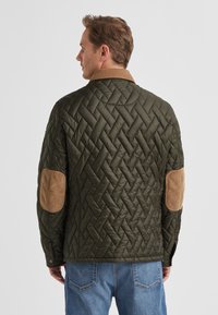 Gesteppte grüne Jacke mit braunen Cord-Ellenbogenpatches und Kragen, mit strukturierter Oberfläche und Druckknopfverschluss am Saum.