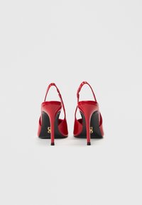 Κόκκινα ψηλοτάκουνά παπούτσια από δερμάτινο βερνίκι με σχέδιο slingback, μυτερή μύτη και μαύρη σόλα με μικρή χρυσή λεπτομέρεια λογότυπου.