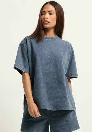 Kobieta o prostych brązowych włosach, ubrana w luźny, wyblakły granatowy T-shirt i pasujące szorty, stojąca na tle jednolitego, jasnego tła.