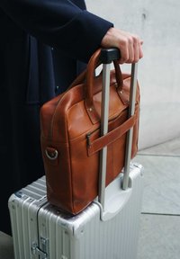 Brown leather briefcase with zippered pocket, carried on the handle of a silver luggage case, showing smooth textures and sturdy design.