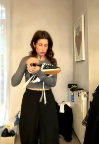 Žena v sivej košeli s dlhými rukávmi a čiernych nohaviciach drží čiernu tenisku s bielymi šnúrkami v svetlej izbe s radiátorom a nástenným umením.